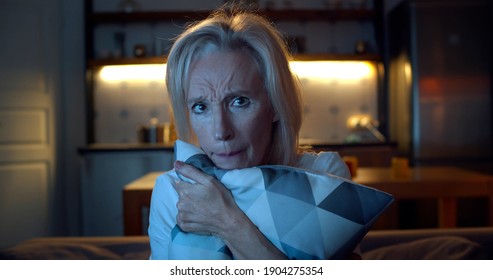 Close Up Of Aged Woman Feeling Scared Watching Horror Movie On Tv And Hugging Pillow. Portrait Of Senior Lady Sitting On Couch In Dark Room Alone And Watching Thriller Film On Tv