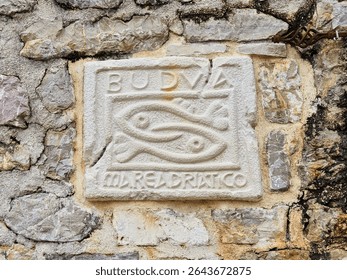 Close up of an ancient bas relief embedded in a stone wall, depicting two fish and bearing the inscription budva mare adriatico, located in budva, montenegro