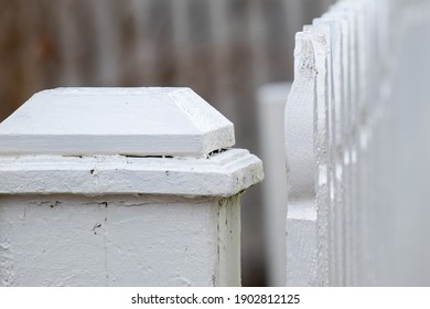 A Close Up Of An Antique White Wooden Picket Fence Post With Vertical Wooden Pailings Having A Curved Shape On Top. The Top Of The Country Barricade Post Has A Square Wood Cap. The Paint Is Worn.