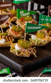 A Close Up Of Appetizers Made Of Nacho Scoops Filled With Guacamole, Sour Cream, Cheese And Salsa And Topped With A Jalapeno On A Dark Wooden Platter.