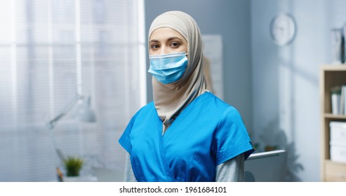 Close Up Of Arabic Young Beautiful Woman General Practitioner In Medical Mask And Uniform Standing In Hospital Looking At Camera. Female Pretty Nurse Working During Covid-19 Pandemic