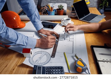Close up of architect hands using divider compass and metal ruler to draw floor plan on blueprint at office desk with helmet.