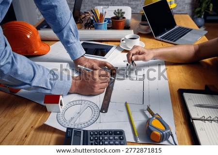 Close up of architect hands using divider compass and metal ruler to draw floor plan on blueprint at office desk with helmet.
