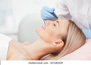 Close Up Of Arms Of Experienced Beautician Undergoing Laser Skin Treatment Of Skin On Female Face. The Young Woman Is Lying And Smiling
