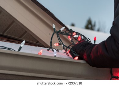 Close up of arms hanging a tangled pile of red and white Christmas lights on a snow covered roof.
