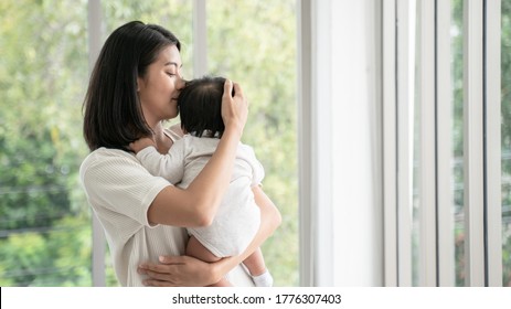 Close up of asian beautiful mom hugging sleeping newborn baby in her arm and kissing the kid.Asian mother look out the window and holding her baby head in home.she is happy to stay at home with baby.