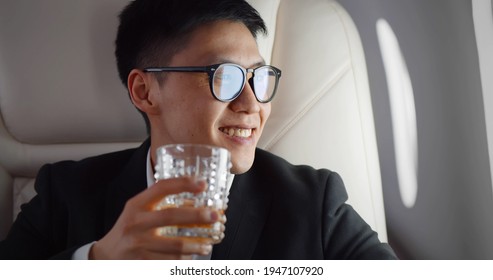 Close Up Of Asian Businessman Drinking Whiskey In Glass In Plane. Portrait Of Korean Rich Man Enjoying Scotch And Looking Out Of Window In Private Jet