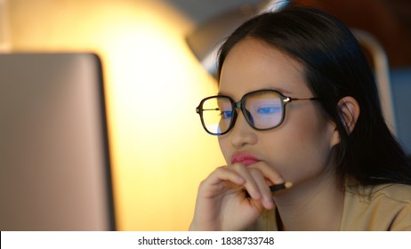 Close Up Of Asian Businesswoman Wearing Glasses Looking At Pc Screen Using A Desktop Computer Working At Night In The Office. Business Graph And Chart Reflection In Her Eyeglasses. Overworked Concept