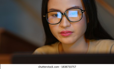 Close Up Of Asian Businesswoman Wearing Glasses Looking At Pc Screen Using A Desktop Computer Working At Night In The Office. Business Graph And Chart Reflection In Her Eyeglasses. Overworked Concept