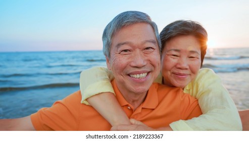 Close Up Of Asian Elderly Carefree Couple Standing On The Beach And Smiling At You Happily In Sunset Background