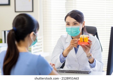 Close Up Of Asian Female Dentist Introduces Patient For Dental Implant By Denture Model While They Wear Protective Face Mask To Prevent COVID19