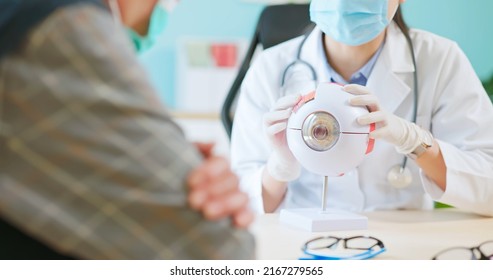 close up of asian female doctor wearing face mask is showing eyeball model and explaining about eyes disease to elder senior man patient in hospital