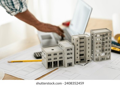 Close up Asian individual architect or engineer holding building scale model of multi-story building. design or review of architectural plans laid out on work surface in front of them. - Powered by Shutterstock