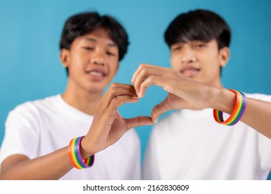  Close Up Of Asian Male Couple With Gay Pride Rainbow Awareness Wristbands Showing Heart Gesture In Studio On Blue Background. Lgbt, Same-sex Love And Homosexual Relationships Concept