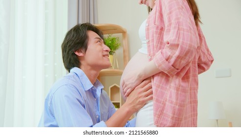 Close Up Of Asian Man Is Kissing Pregnant Wife Belly And Expecting Their Baby Coming Near Window At Home