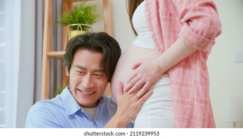 Close Up Of Asian Man Is Listening Pregnant Wife Belly And Expecting Their Baby Coming Near Window At Home