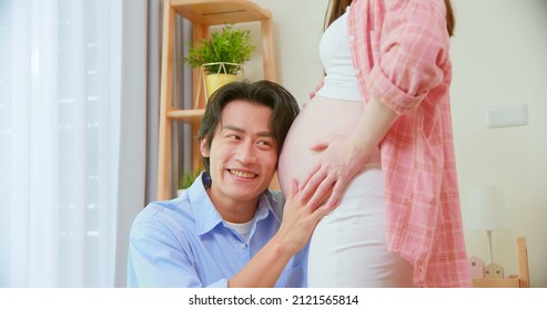 Close Up Of Asian Man Is Listening Pregnant Wife Belly And Expecting Their Baby Coming Near Window At Home