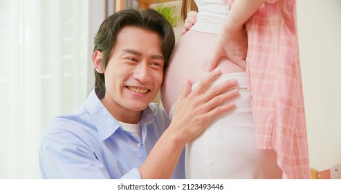 Close Up Of Asian Man Is Listening Pregnant Wife Belly And Expecting Their Baby Coming Near Window At Home