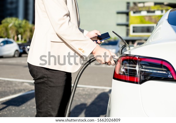 close up of asian man use smart phone while waiting\
and power supply connect to electric vehicles for charging the\
battery in car