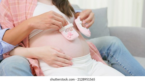 close up of asian man is using baby shoes touching pregnant wife belly and expecting their child coming on couch at home