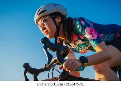 close up asian woman with athletic body shape in protective helmet and glasses riding bicycle on blue sky 
