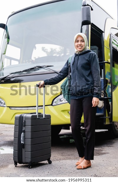 close up of asian woman in a veil smiles looking at\
the camera while holding a back suitcase against the background of\
the bus
