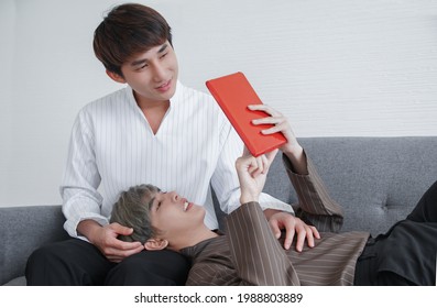 Close Up Of Asian Young Gay Couple Looking At Tablet Together On Sofa At Home. A Man Touch His Lover's Hairs With Love Who Lying On His Boyfriend Lap.  LGBT Gay Couple Relaxing Lifestyle Concept.