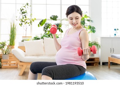 Close Up Of Asian Young Pregnant Woman Doing Exercise With Pilates Ball And Dumbbells In Living Room