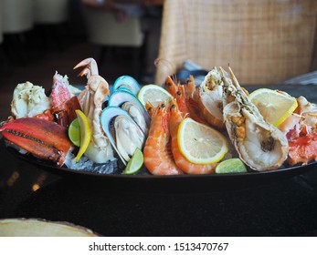 Close Up Assorted Fresh Cooked Cold Seafood As Shrimps, Prawns, New Zealand Mussels, Mussels, Oyster, Rock Lobster, Lobster Claw With Yello Leomon In Oval Shaped Dish Or Plate On Ice On Wooden Table