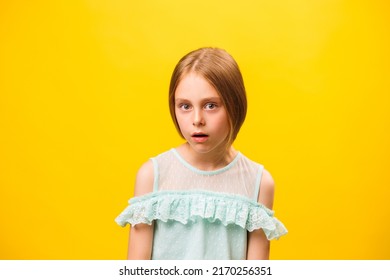 Close Up Of An Astonished Wow Little Girl Child Looking At Camera. Kid With Open Mouth He Is Very Excited And Touches His Face. Cropped Studio Shot With Yellow Background