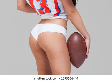 Close Up Of The Athletic Buttocks Of A Girl Dressed In A Jersey T-shirt And Holding An American Football Ball Isolated On Grey Background