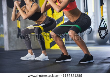Close up of athletic women in squat together in gym. Couple of fit girls are exercising with resistance band for lower body relief. They are wearing sport clothes and sneakers