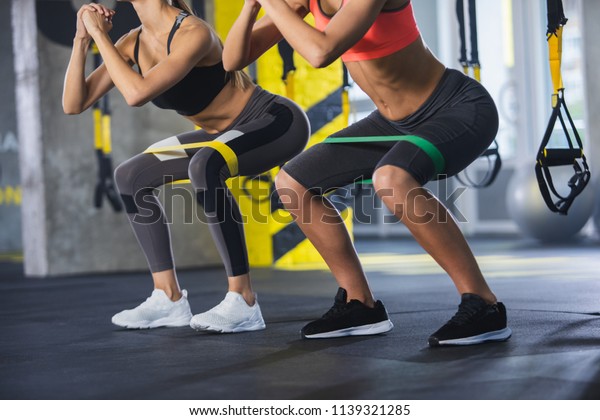 Close up of athletic\
women in squat together in gym. Couple of fit girls are exercising\
with resistance band for lower body relief. They are wearing sport\
clothes and sneakers