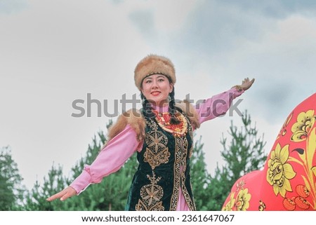 Close up of attractive smiling Asian girl in fur headdress and traditional festive Bashkir clothes dancing bird dance against background of sky. Village festival, Siberia, Russia.Copy space