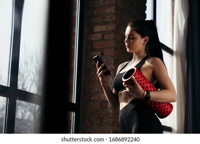 Close up attractive sporty girl with smartphone is standing in front of a panoramic window and holds a red cylinder. - Powered by Shutterstock