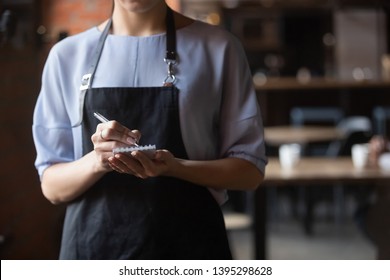 Close up attractive waitress wearing black apron standing in cozy coffeehouse, female cafe worker with notebook in hands waiting for, ready to take customers, guests order in restaurant