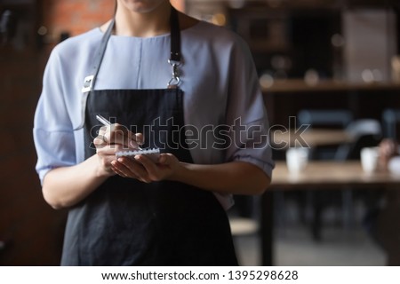 Close up attractive waitress wearing black apron standing in cozy coffeehouse, female cafe worker with notebook in hands waiting for, ready to take customers, guests order in restaurant