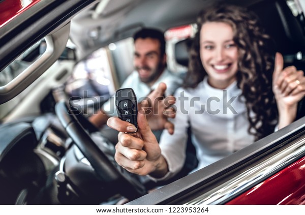 Close up of attractive woman showing car key through\
the window. Nice young couple sitting inside the new car at the\
dealership. Focus on key