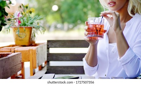 Close up of an attractive young blonde woman in white shirt drinking a cocktail through a straw, sitting on veranda of modern restaurant on a sunny summer day.