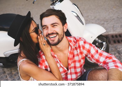 Close up of attractive young couple whipsering to ear a secret while sitting near scooter and smiling. Happy relathionship concept