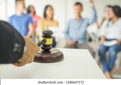 Close up auctioneer hand, black suite, holding gavel, wooden hammer, and blur group of people in auction room, one man raising hand up for bidding. Product or project auction market concept background