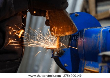 Close up of axe blade being sharpened on bench grinder, metal edge pressed to rotating abrasive wheel with bright sparks in workshop during tool repair.