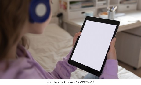 Close Up Back Rear View Little Teen Girl In Headphones Holding Digital Computer Tablet With White Mock Up Screen In Hands, Educational Application For Children Or Educational Software Advertisement.