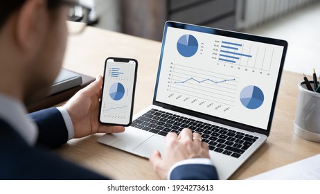 Close up back view of businessman work on computer and cellphone consider financial diagrams graphs. Male CEO or employee analyze finances statistics on laptop and smartphone. Banking concept. - Shutterstock ID 1924273613