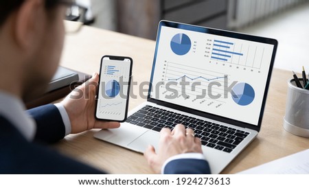 Close up back view of businessman work on computer and cellphone consider financial diagrams graphs. Male CEO or employee analyze finances statistics on laptop and smartphone. Banking concept.