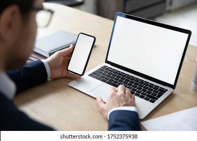 Close up back view of male employer sit at desk using modern laptop and smartphone gadget, businessman consult client work on computer and cellphone device at workplace, white mockup screen