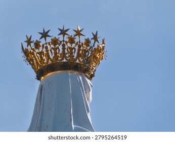 Close up back view of the white statue of Our Lady of Lourdes featuring her iconic golden crown with stars against a clear blue sky background. Sanctuary of Our Lady of Lourdes, France.