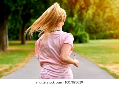 Close Up From The Back Of A Young Blonde Woman Running Away From The Camera On The Road In A Park
