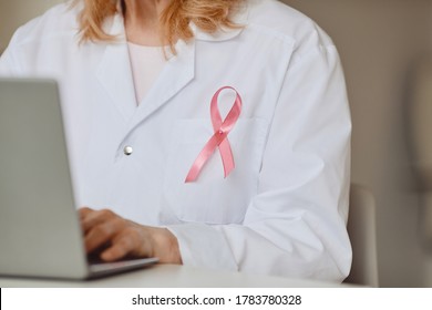 Close Up Background Of Pink Ribbon Pinned On White Lab Coat Of Female Doctor Using Laptop, Breast Cancer Awareness Symbol, Copy Space