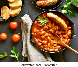 Close Up Of Baked White Beans In Tomato Sauce And Grilled Sausages In Frying Pan Over Black Stone Background. Top View, Flat Lay